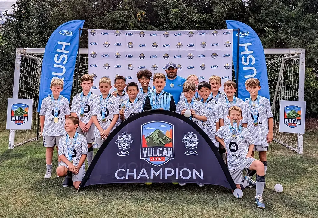 Soccer team poses with championship banner and medals. Players standing in front of a backdrop and goals.