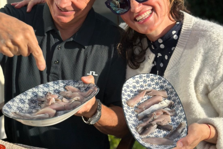 Two people smiling and holding plates of fish outdoors in bright sunlight.