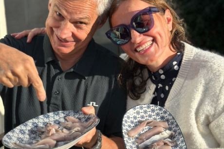 Two people smiling and holding plates of fish outdoors in bright sunlight.