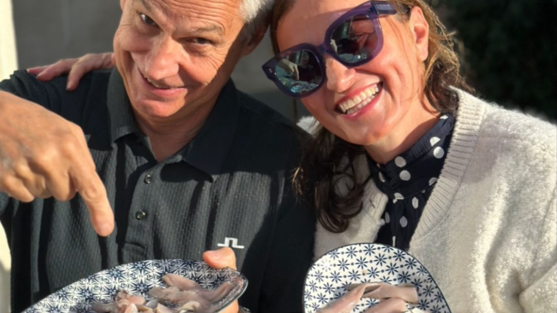 Two smiling people outdoors holding plates of fish at a table