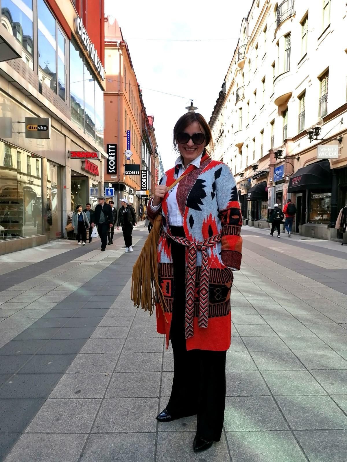Woman in a red-and-white coat standing on a city street lined with shops.