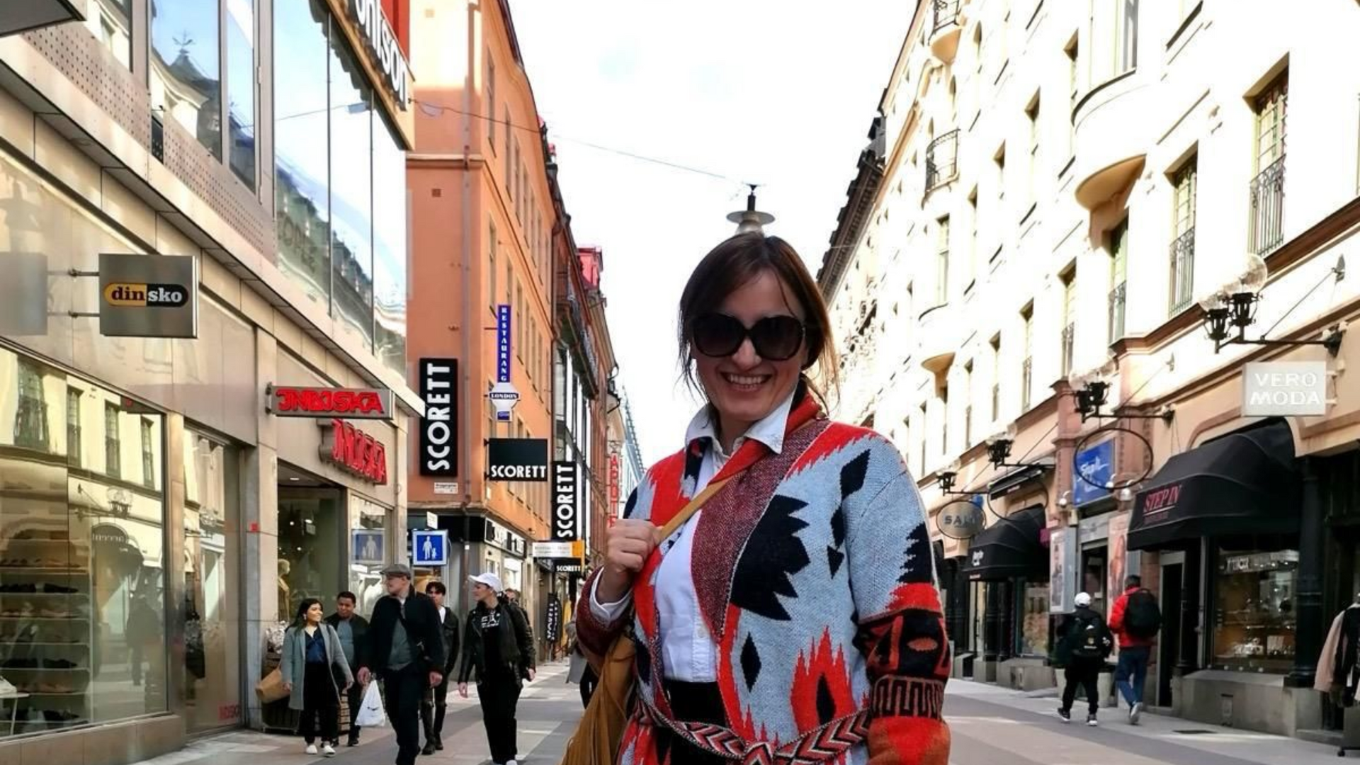 Person in sunglasses and a red patterned coat standing on a busy pedestrian street between tall buildings.