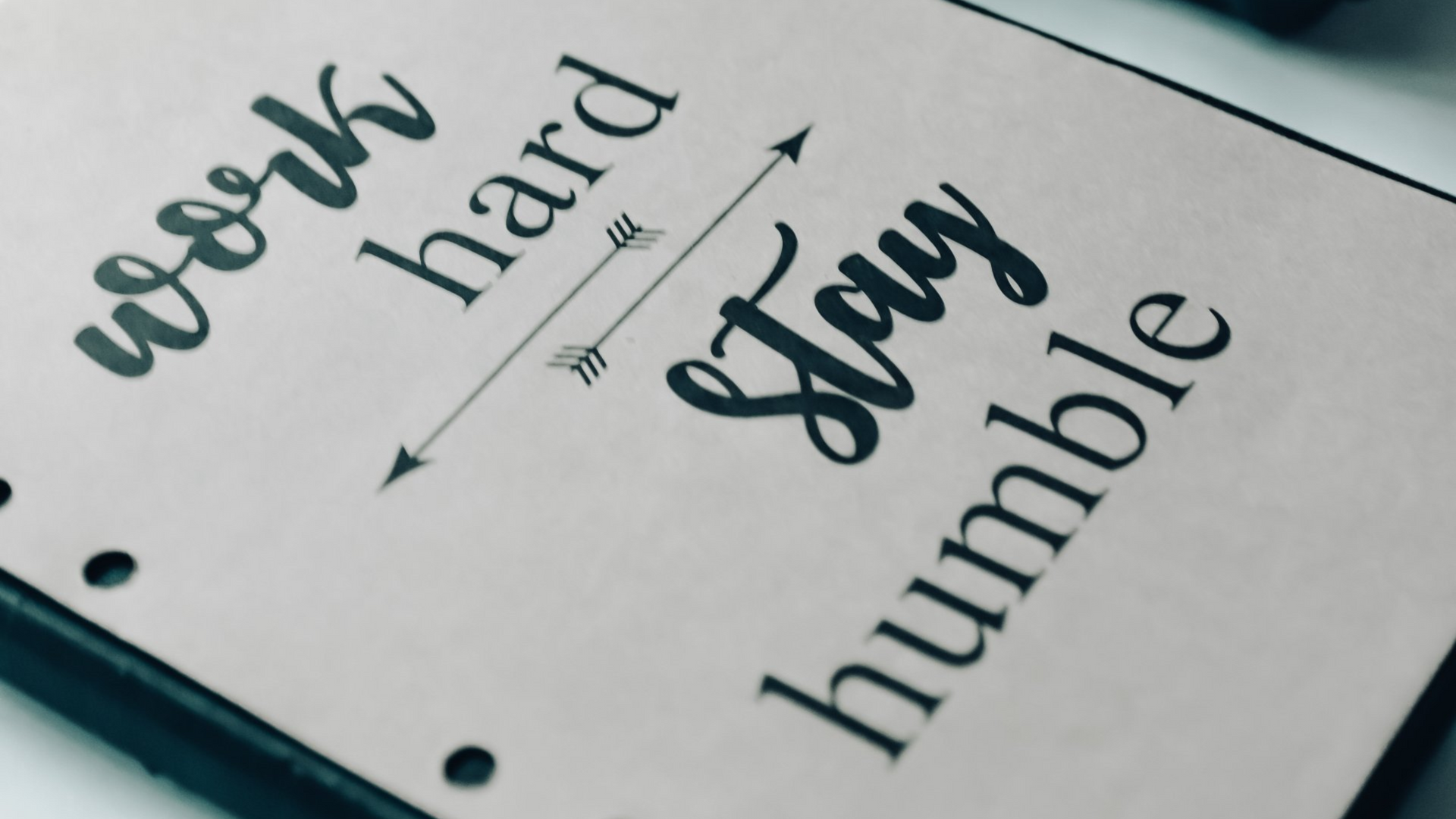 Notebook with motivational quote “work hard, stay humble” beside a camera and laptop on a desk