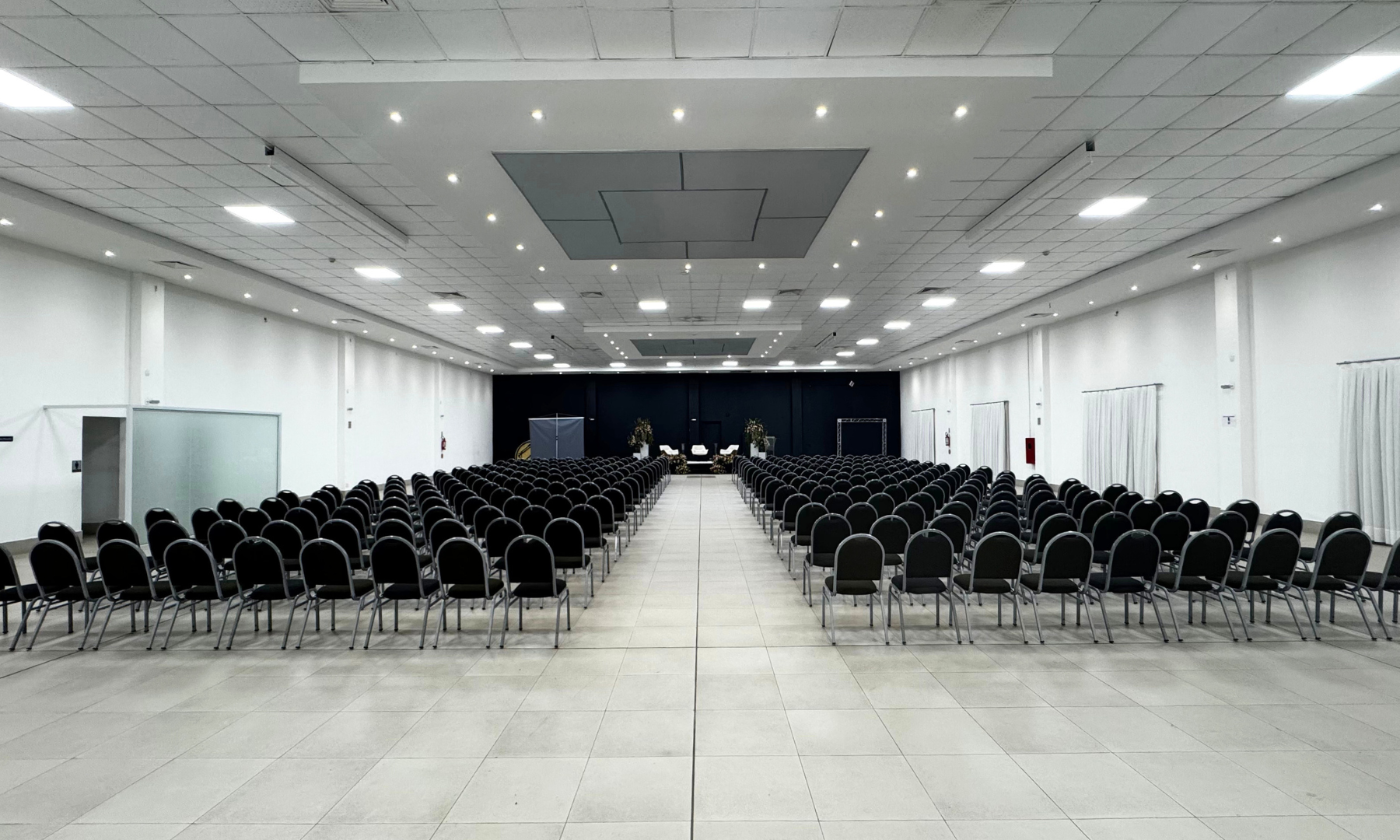 Fileiras de cadeiras pretas estão voltadas para um palco escuro em um amplo espaço para eventos, vazio e com paredes brancas.