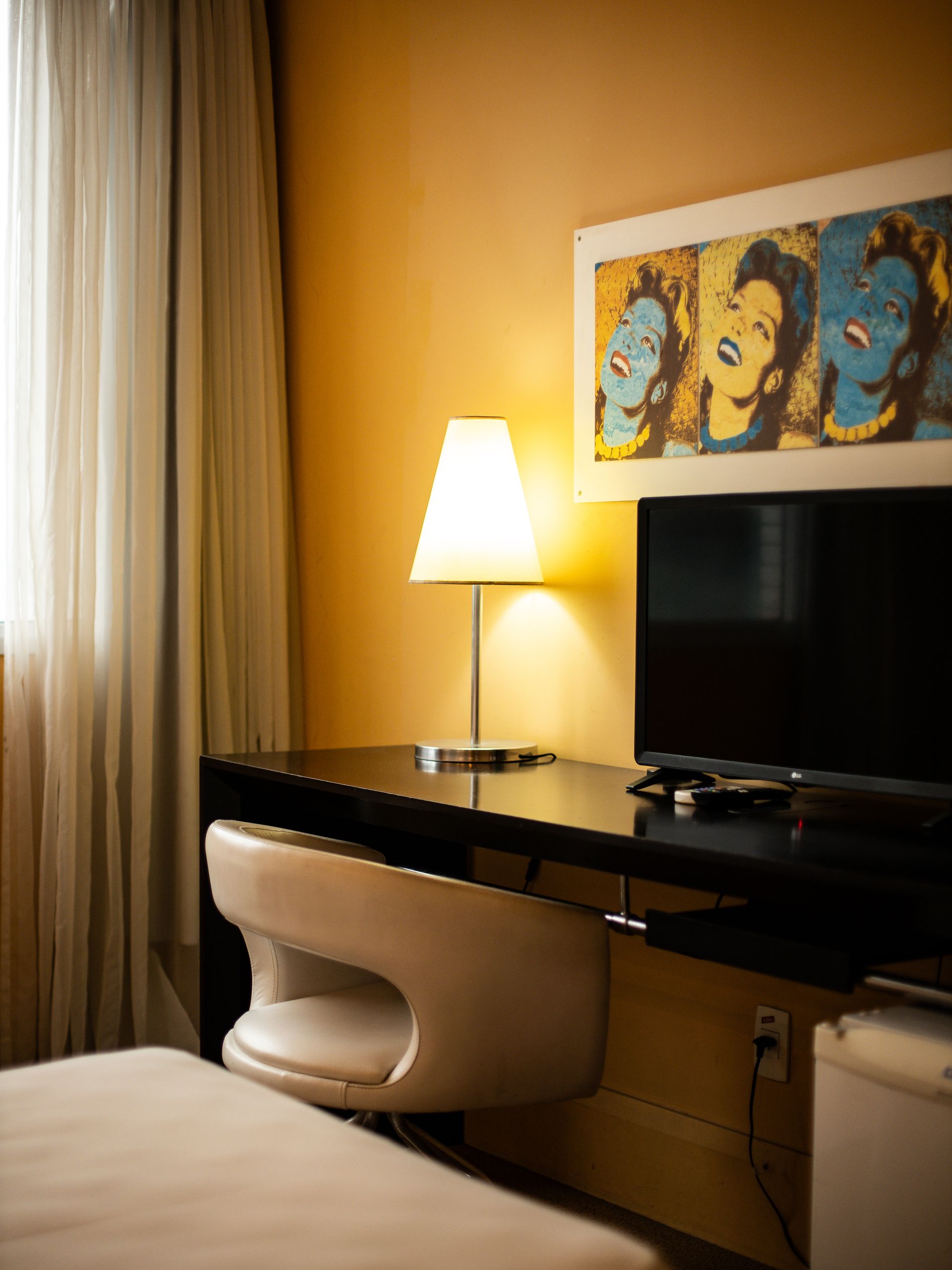 Um quarto de hotel com escrivaninha, cadeira, TV, luminária e obras de arte pop coloridas acima da televisão.