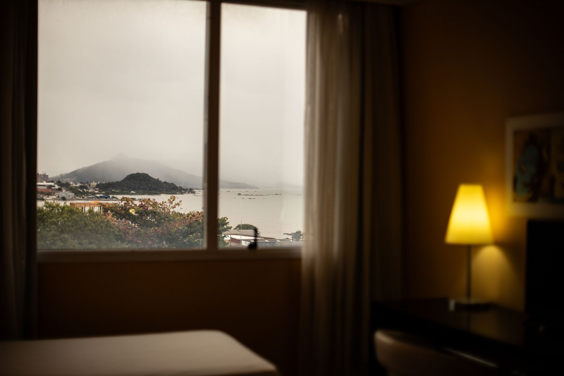 Vista da janela para um mar nublado e uma colina, vista de um quarto com uma lâmpada acesa.