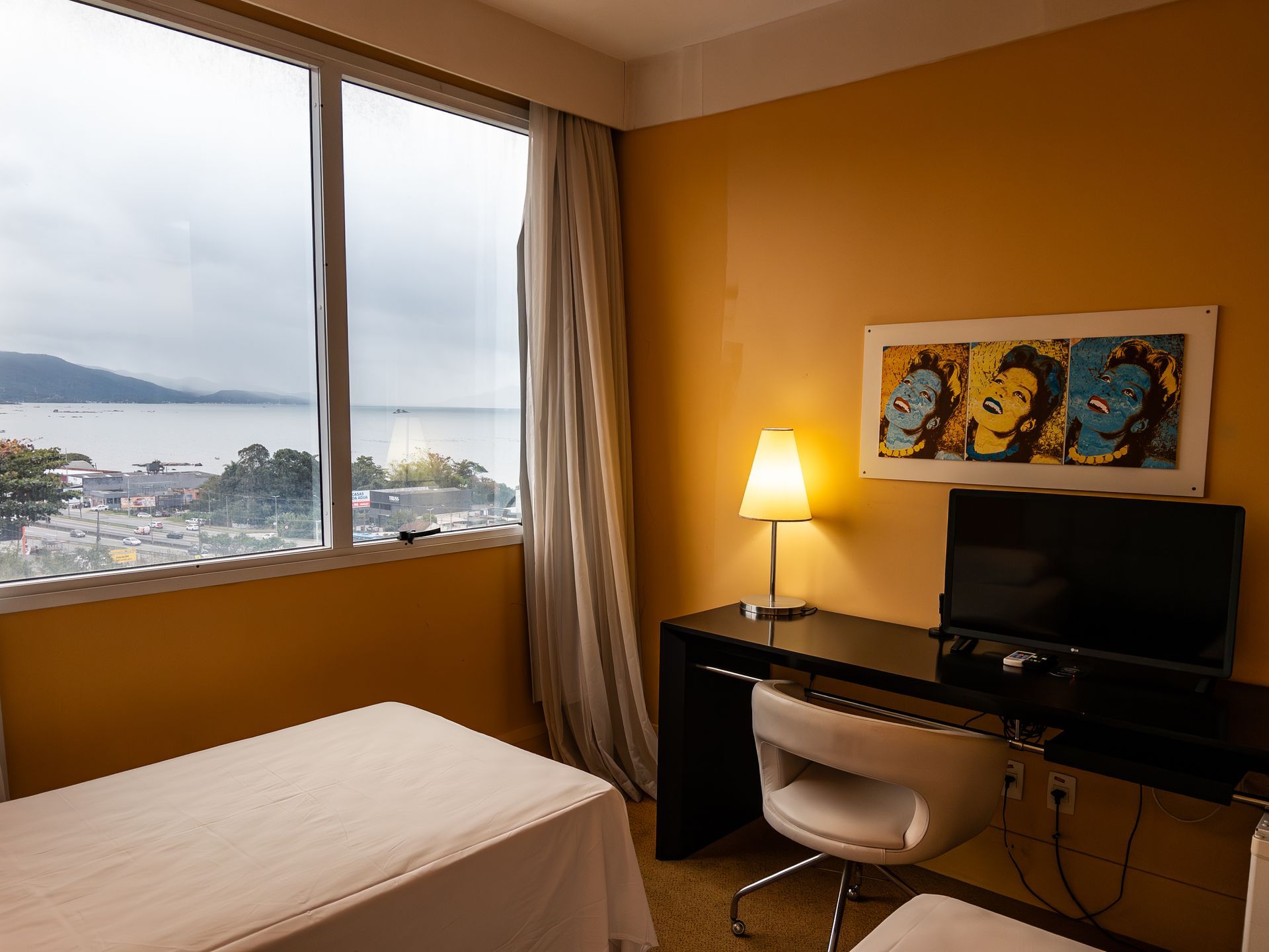 Quarto de hotel com uma grande janela com vista para o litoral. Inclui escrivaninha, cadeira, luminária e obra de arte.