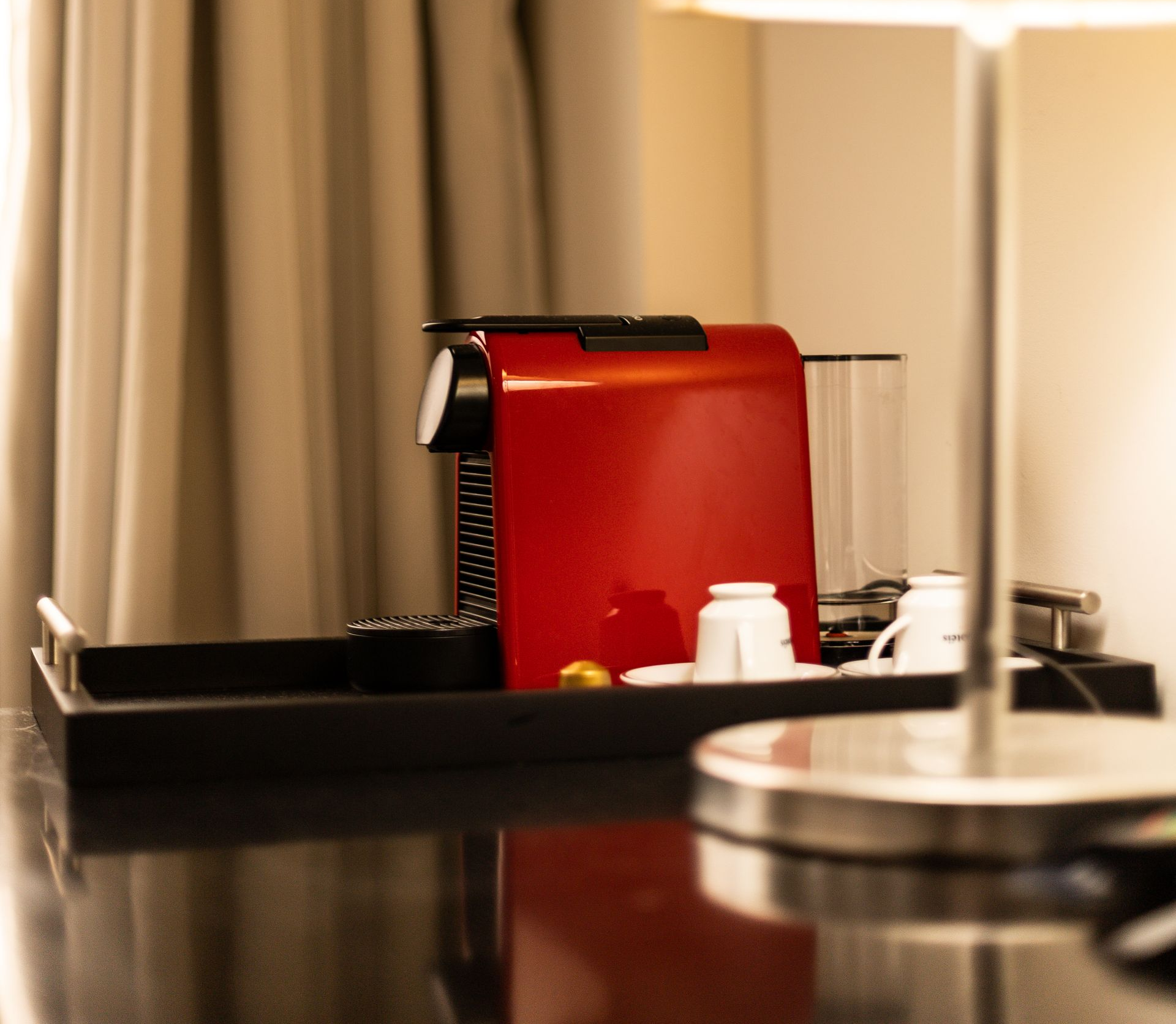 Máquina de café vermelha e xícaras sobre uma bandeja preta, ao lado de um abajur em uma sala com cortinas bege.