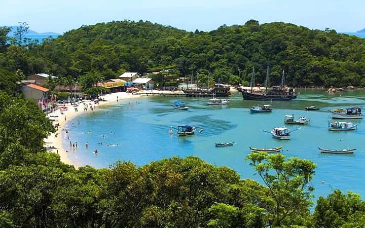 Baía com barcos, praia e colinas verdejantes sob um céu azul brilhante.