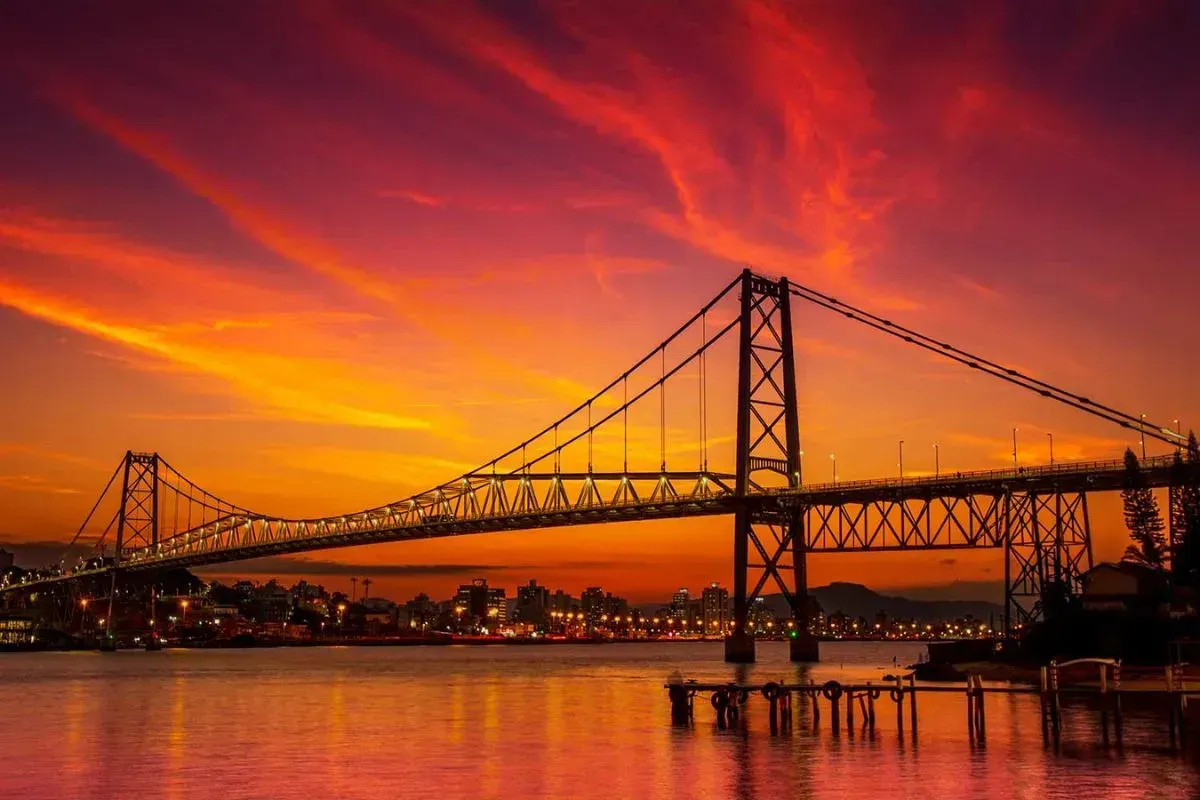 A silhueta da ponte contra um pôr do sol flamejante sobre a água, com o céu em tons de laranja e vermelho.