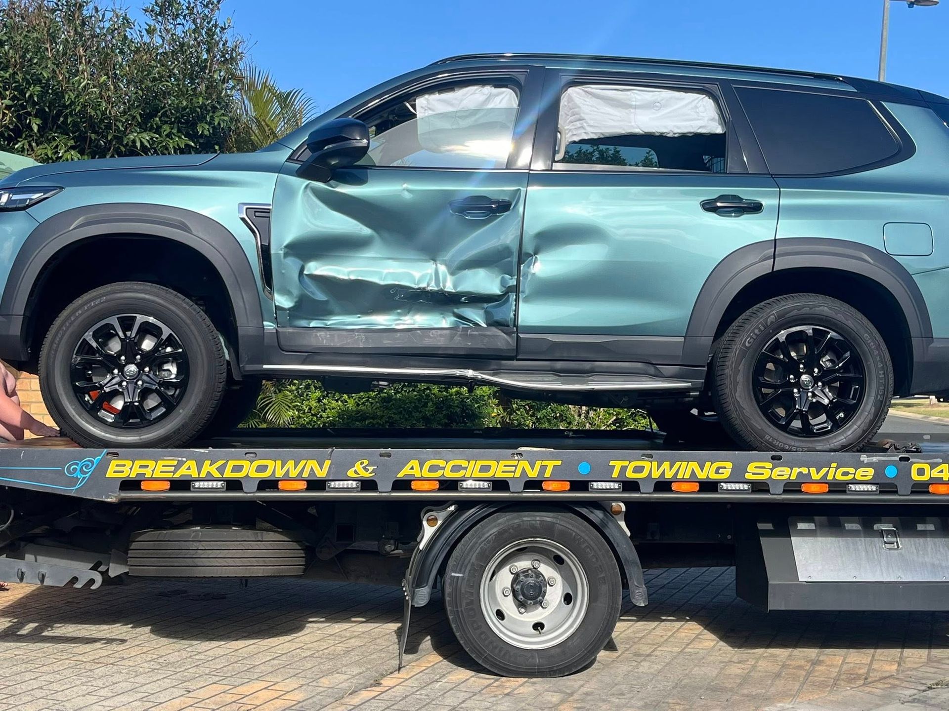 Damaged teal SUV strapped on a tow truck under a clear blue sky — A B & Forge Towing In Narangba, QLD