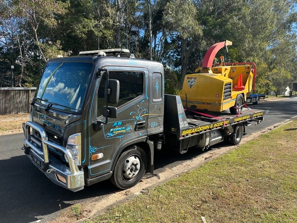 Gray Flatbed Truck Carrying Blue Construction Equipment Parked Outdoors — A B & Forge Towing In Caboolture, QLD
