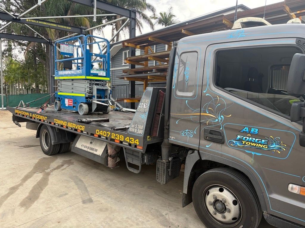 Gray flatbed truck carrying blue construction equipment parked outdoors under a metal structure — A B & Forge Towing In Caboolture, QLD