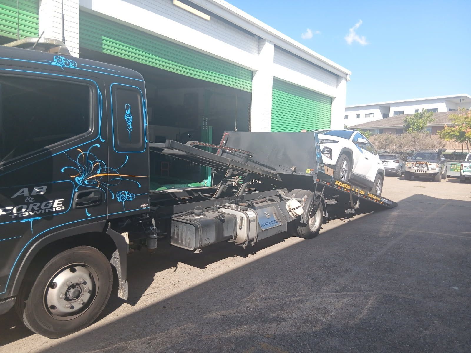 Tow truck hauling a damaged car in front of a green garage on a sunny street — A B & Forge Towing In Narangba, QLD