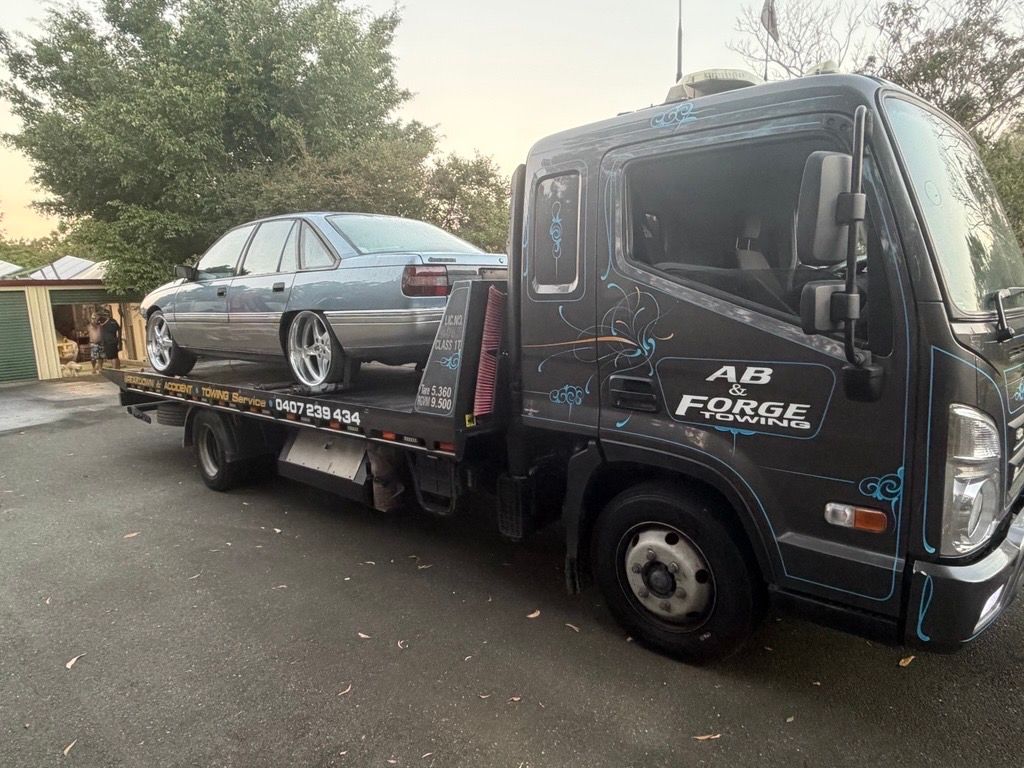 Black tow truck carrying a gray sedan on a flatbed at dusk — A B & Forge Towing In Narangba, QLD