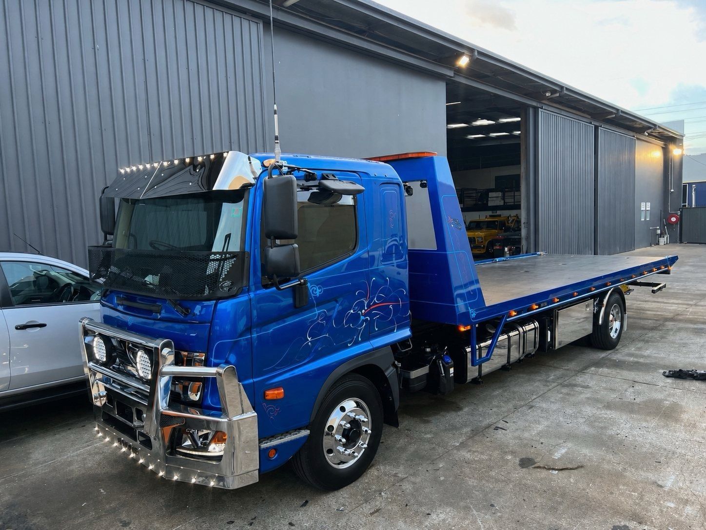 Blue flatbed tow truck parked beside a warehouse on a wet lot — A B & Forge Towing In Caboolture, QLD