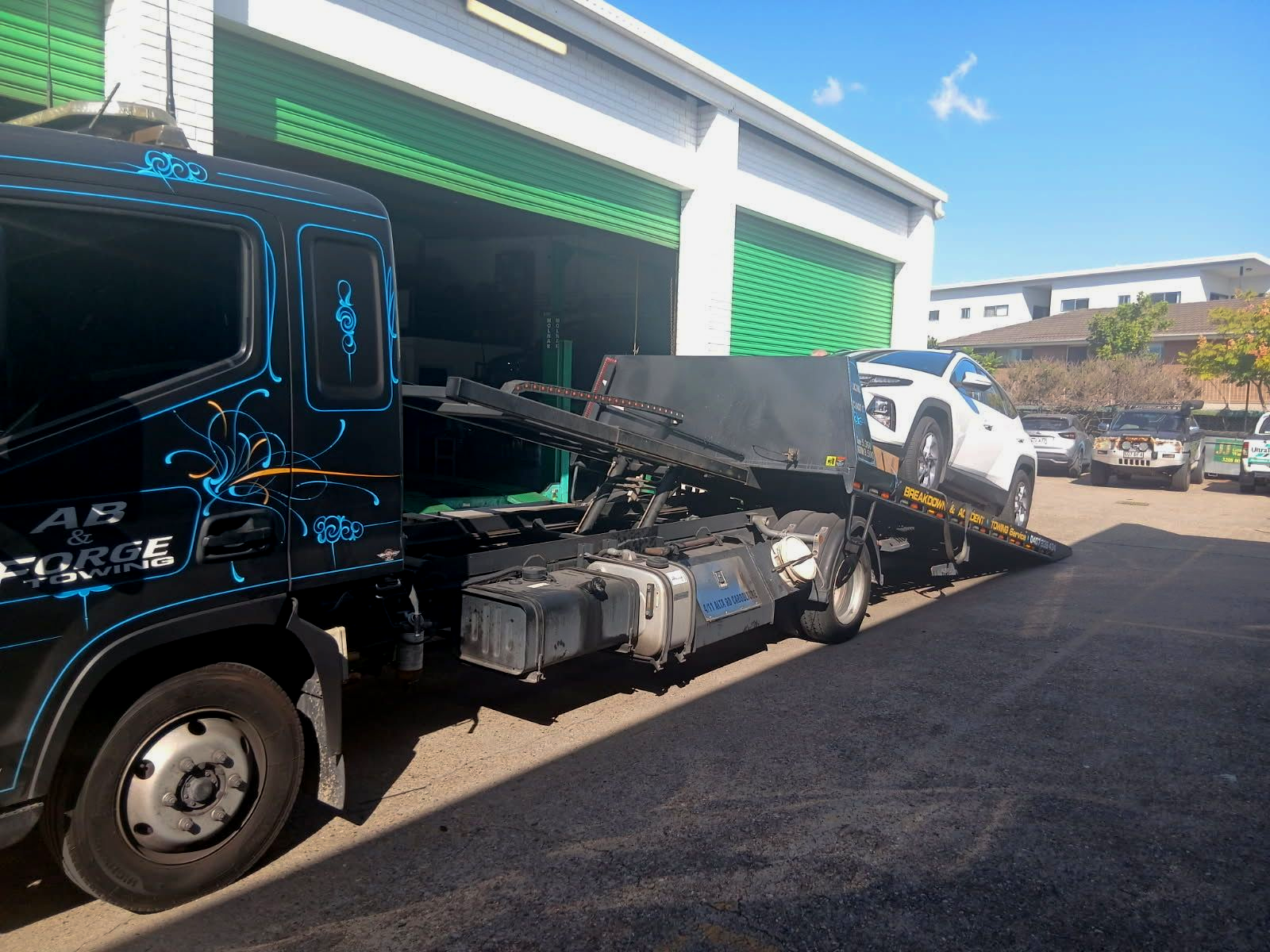 A Black Tow Truck Carries a Wrecked Car Beside a Green Garage — A B & Forge Towing In Caboolture, QLD