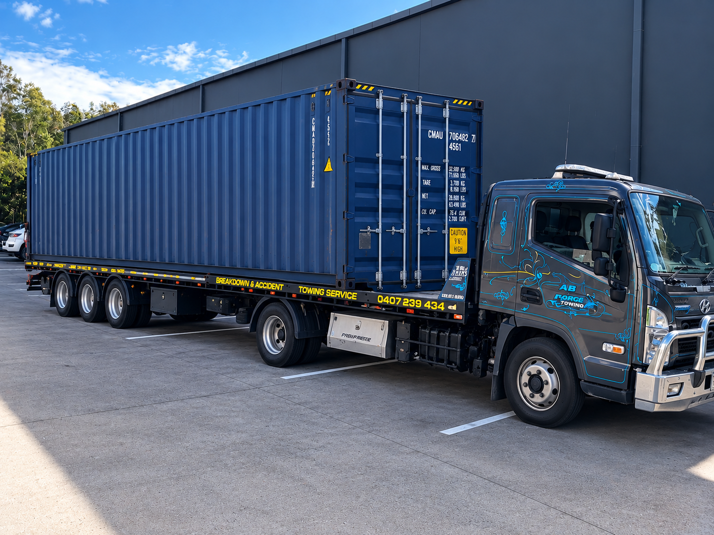 Blue semi-truck with a flatbed trailer carrying a large blue shipping container beside a warehouse — A B & Forge Towing In Caboolture, QLD