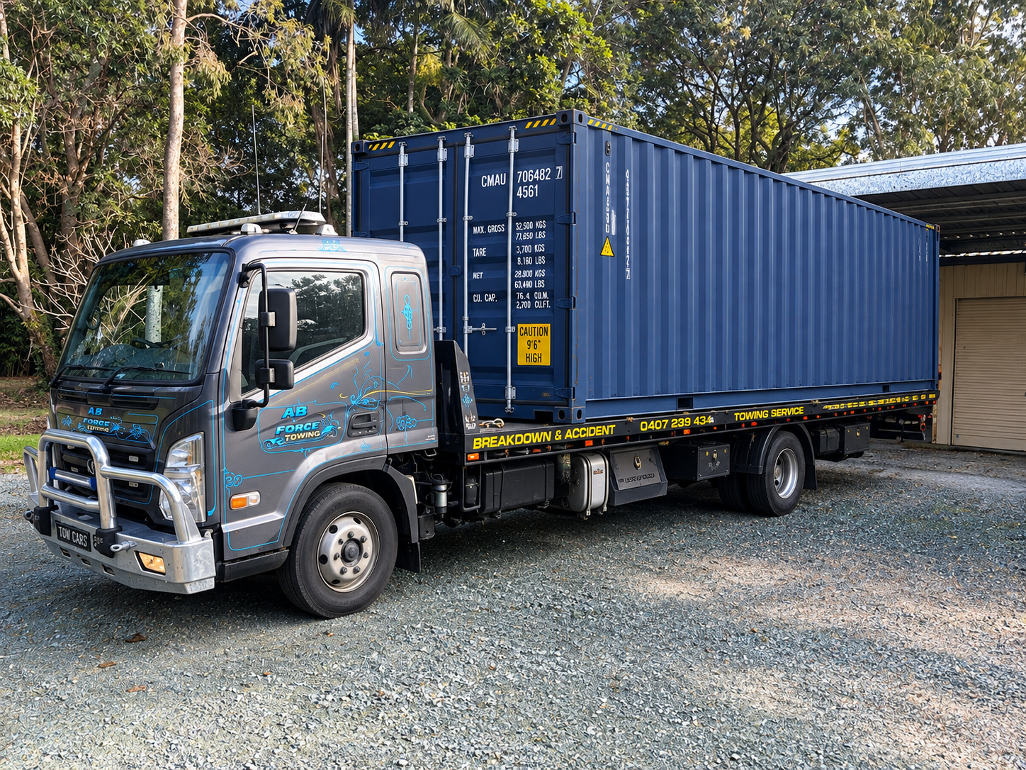 Silver flatbed truck carrying a blue shipping container on gravel near trees and a building — A B & Forge Towing In Caboolture, QLD