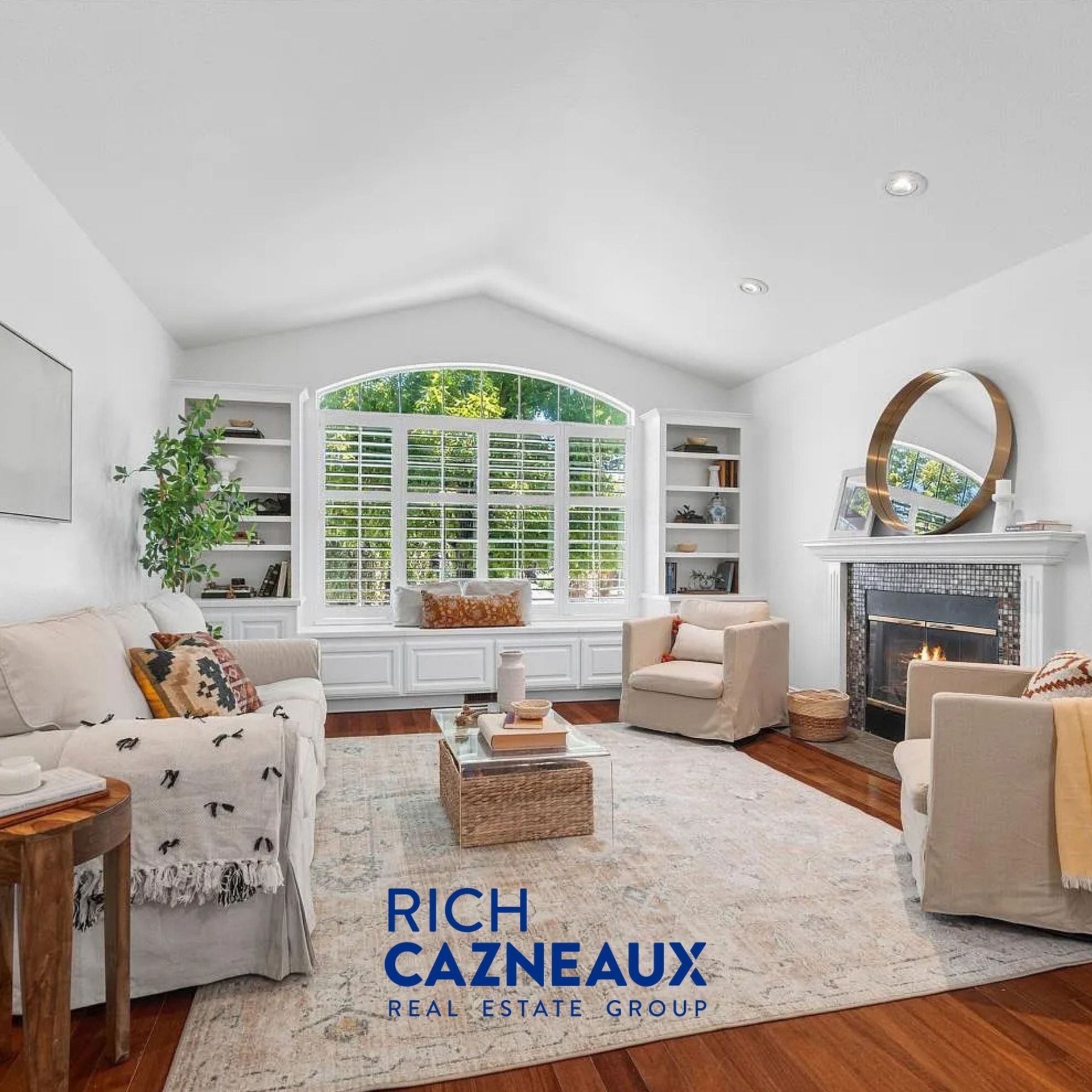 Bright living room with vaulted ceiling, built-ins, fireplace, large arched window, and hardwood floors.