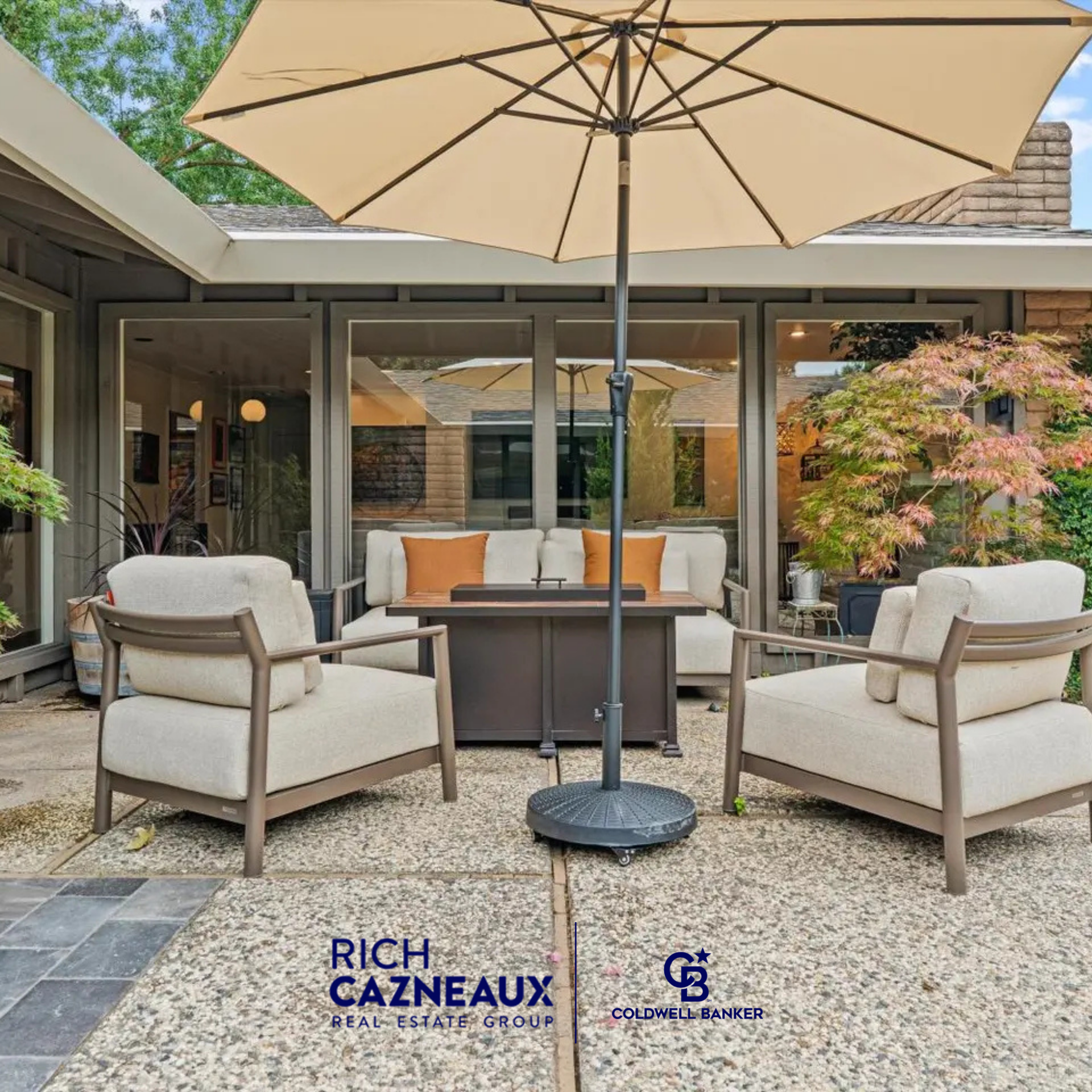 A patio seating area with umbrella, two armchairs, and sofa outside a home with glass doors with Rich Cazneaux branding.