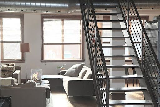 Modern loft-style living area with huge windows, sofas, a sleek staircase, and urban elements, creating a cozy atmosphere.