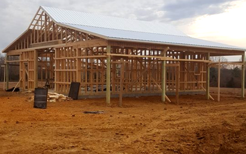 Wooden frame of a barn under construction with a white metal roof, set on a dirt lot against a cloudy sky.