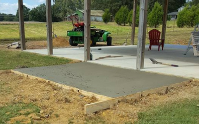 Freshly poured concrete slab extending from a covered patio, with a lawn mower, tools, and red chair in the background.