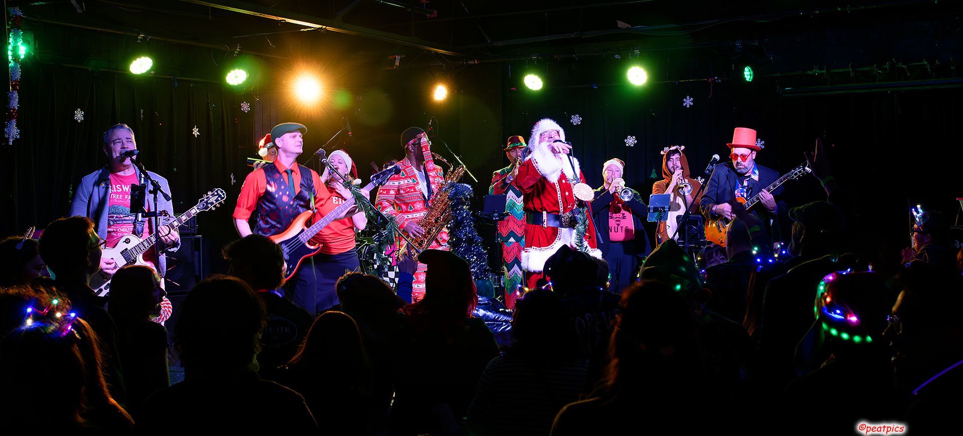 A band performs on a dimly lit stage, with one member dressed as Santa Claus, in front of a dark, silhouetted crowd.