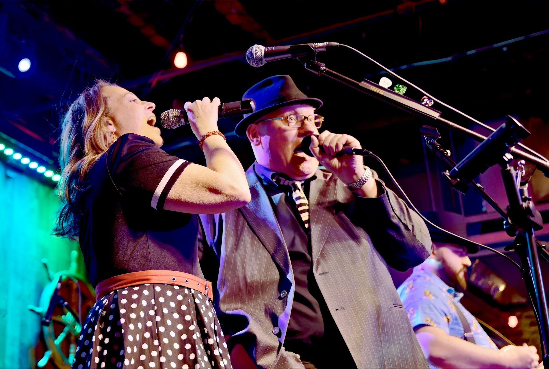 A woman in a polka-dot skirt and a man in a hat and blazer singing together into microphones on a stage.