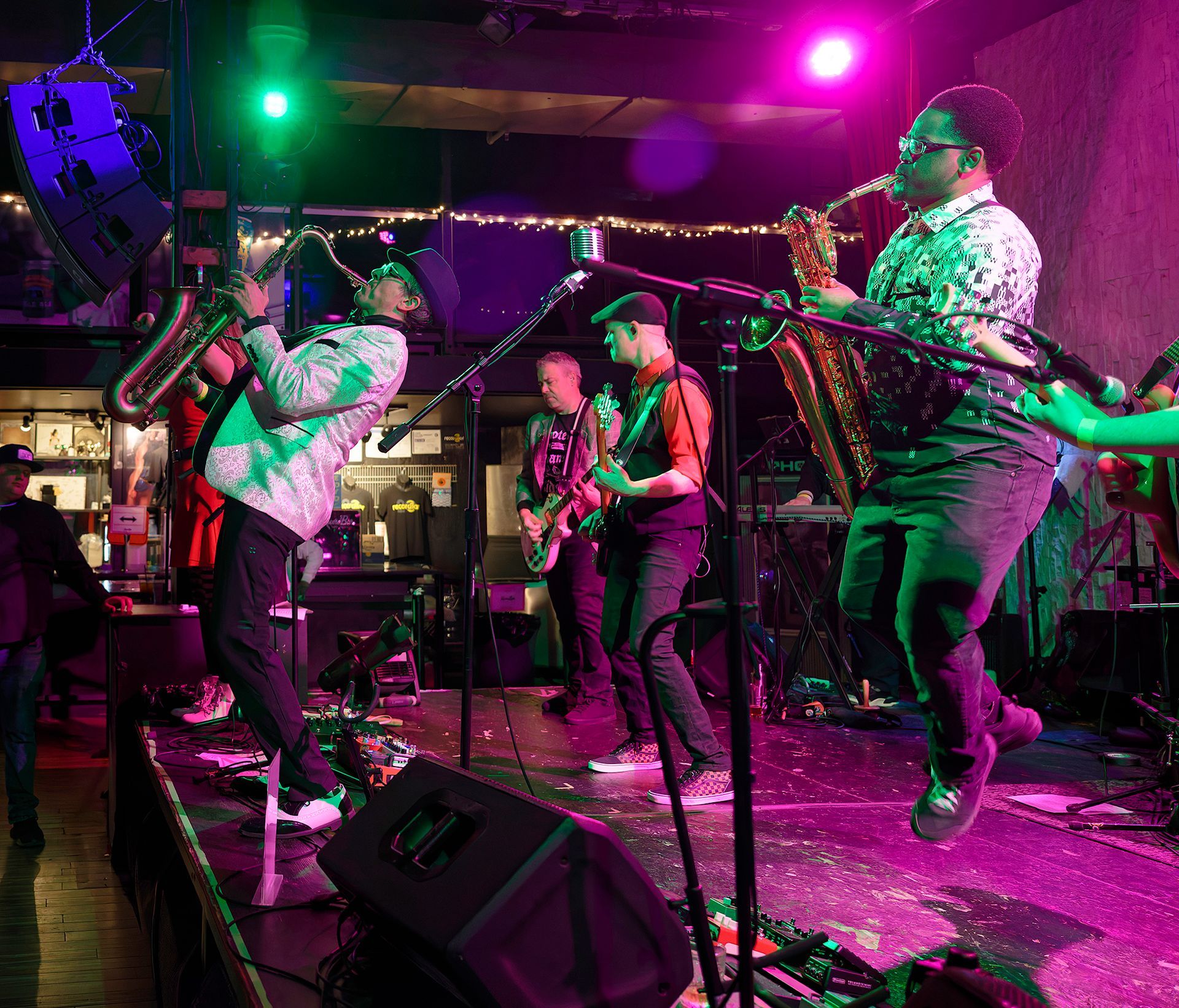 Live band performing on a stage with colorful green and pink lights, guitars, and microphones