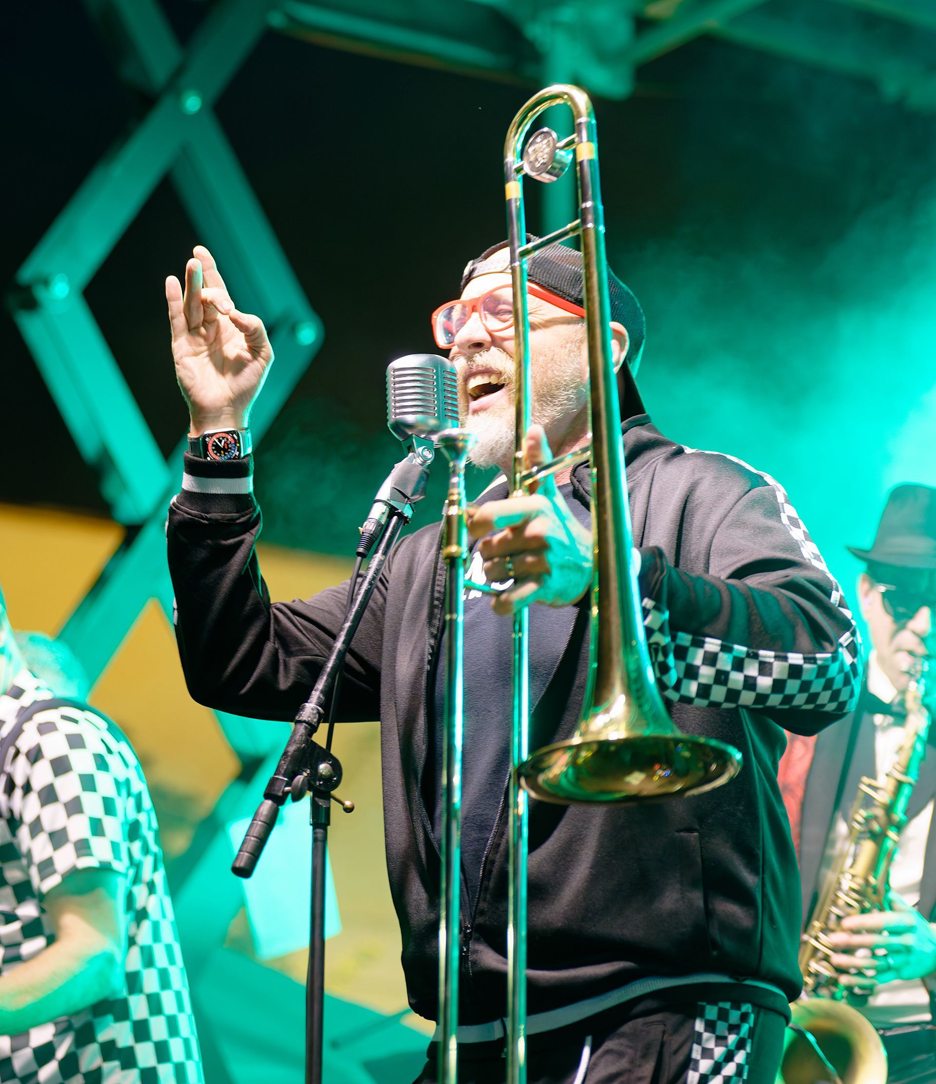 Musician playing trombone on a lit stage, smiling beside a microphone, with green and yellow lighting.