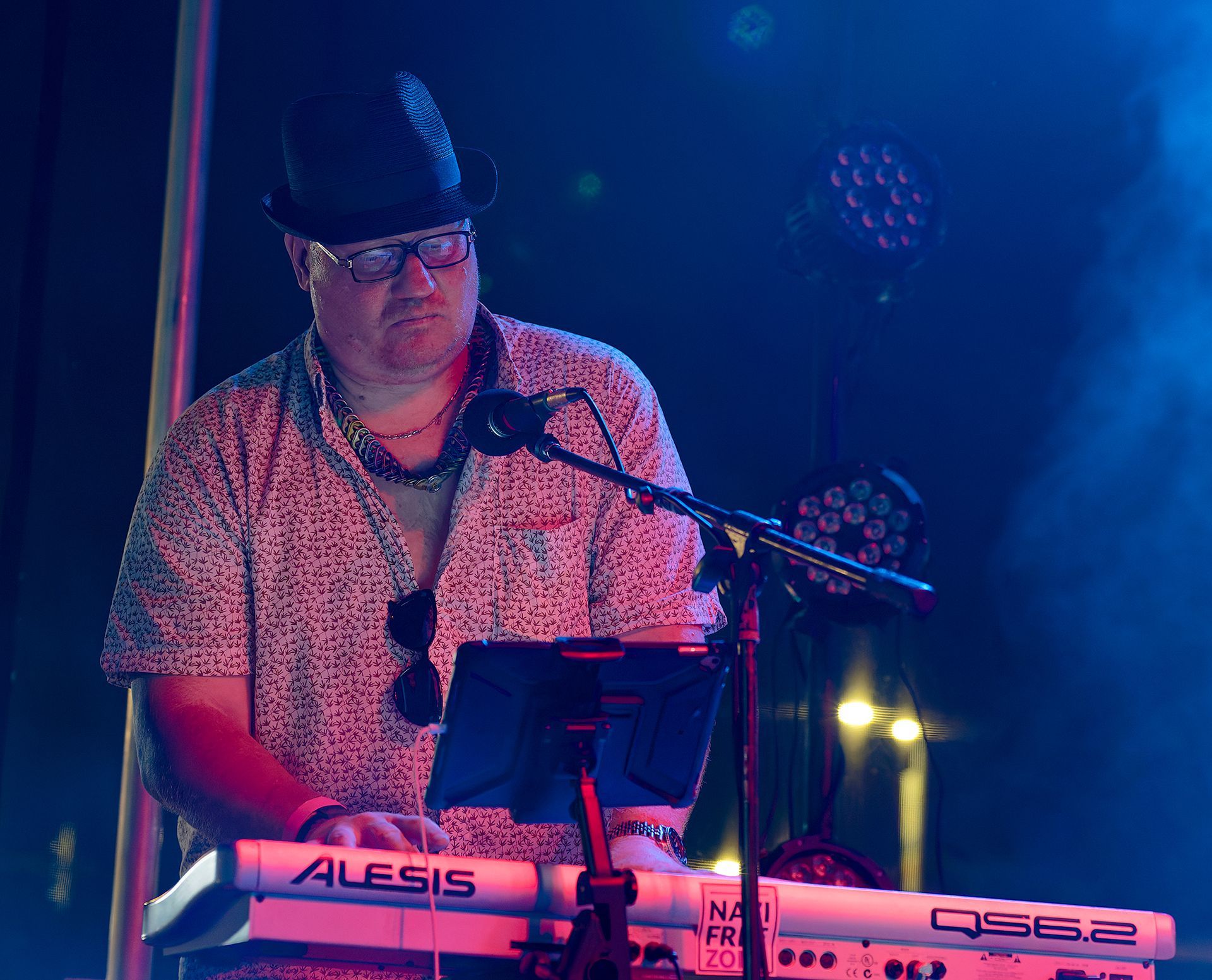Musician playing keyboard on a lit stage, wearing a hat and glasses under purple-blue lights.