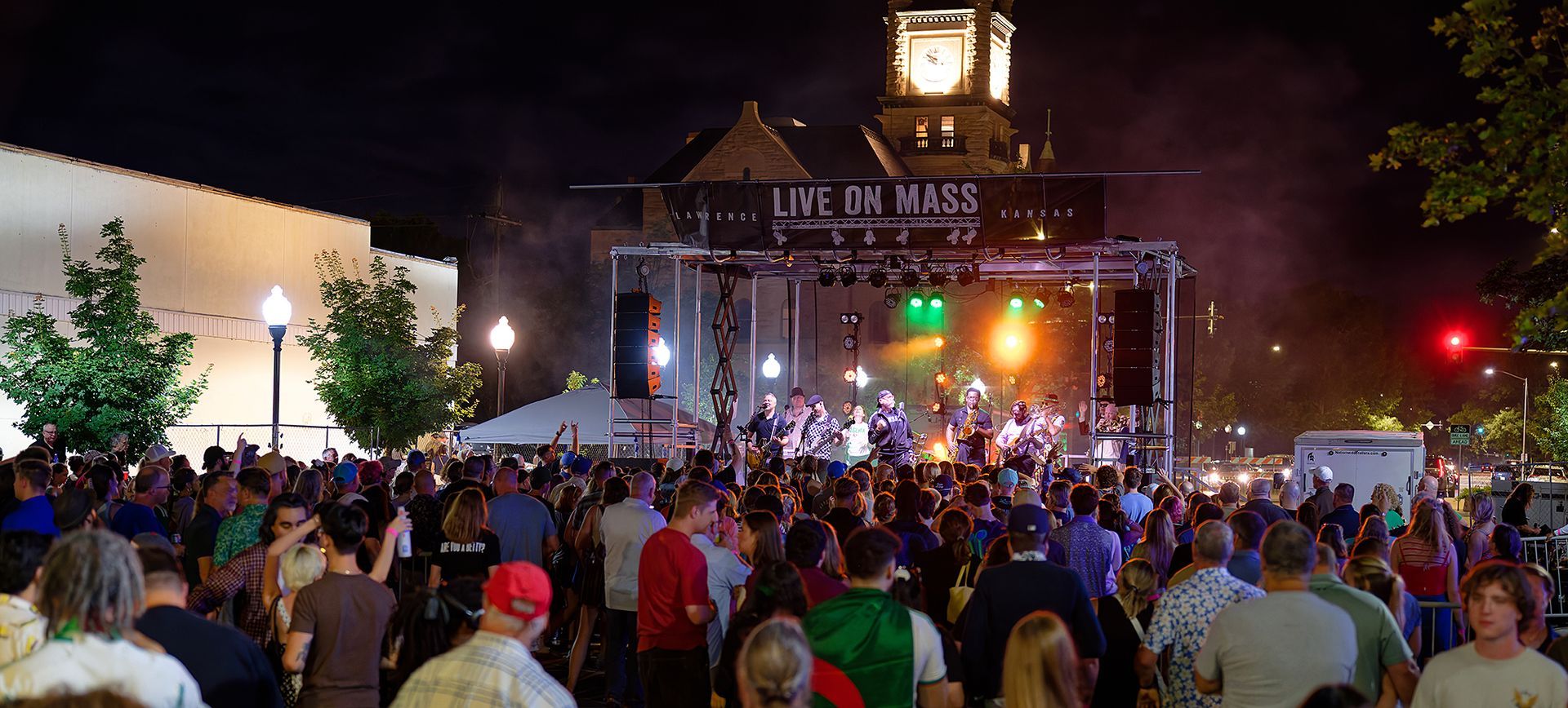 Crowd gathered at a nighttime concert under a lit stage with a clock tower backdrop.