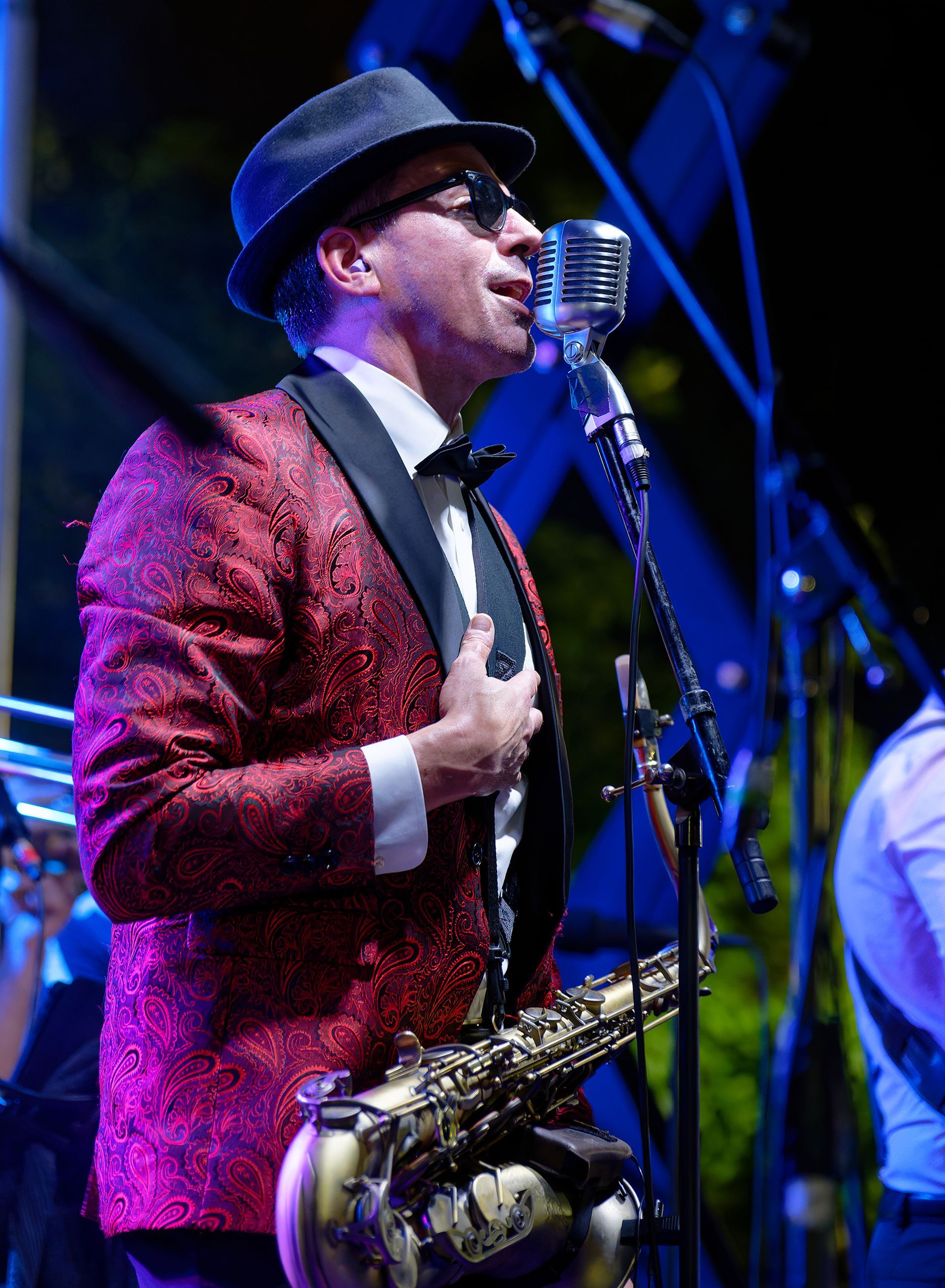 Singer in a red blazer and black hat performing at a vintage microphone with a saxophone on a stand nearby.