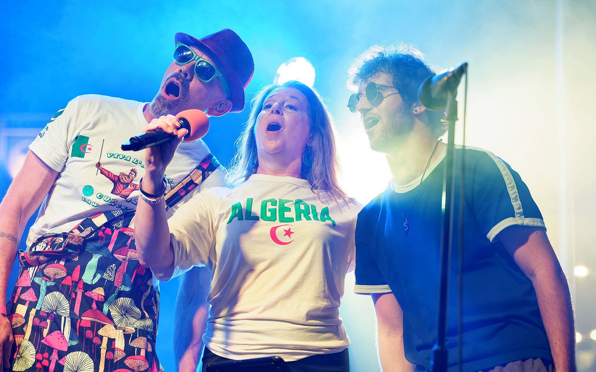Three singers on stage under bright blue and green lights, each holding a microphone and singing.