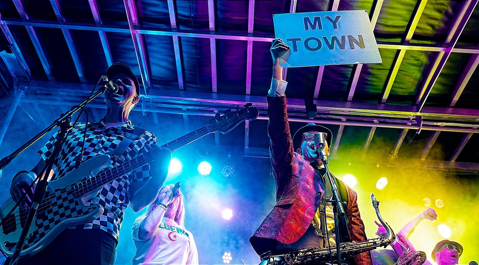 Crowd at concert under blue and yellow lights, one person holding a “MY TOWN” sign overhead