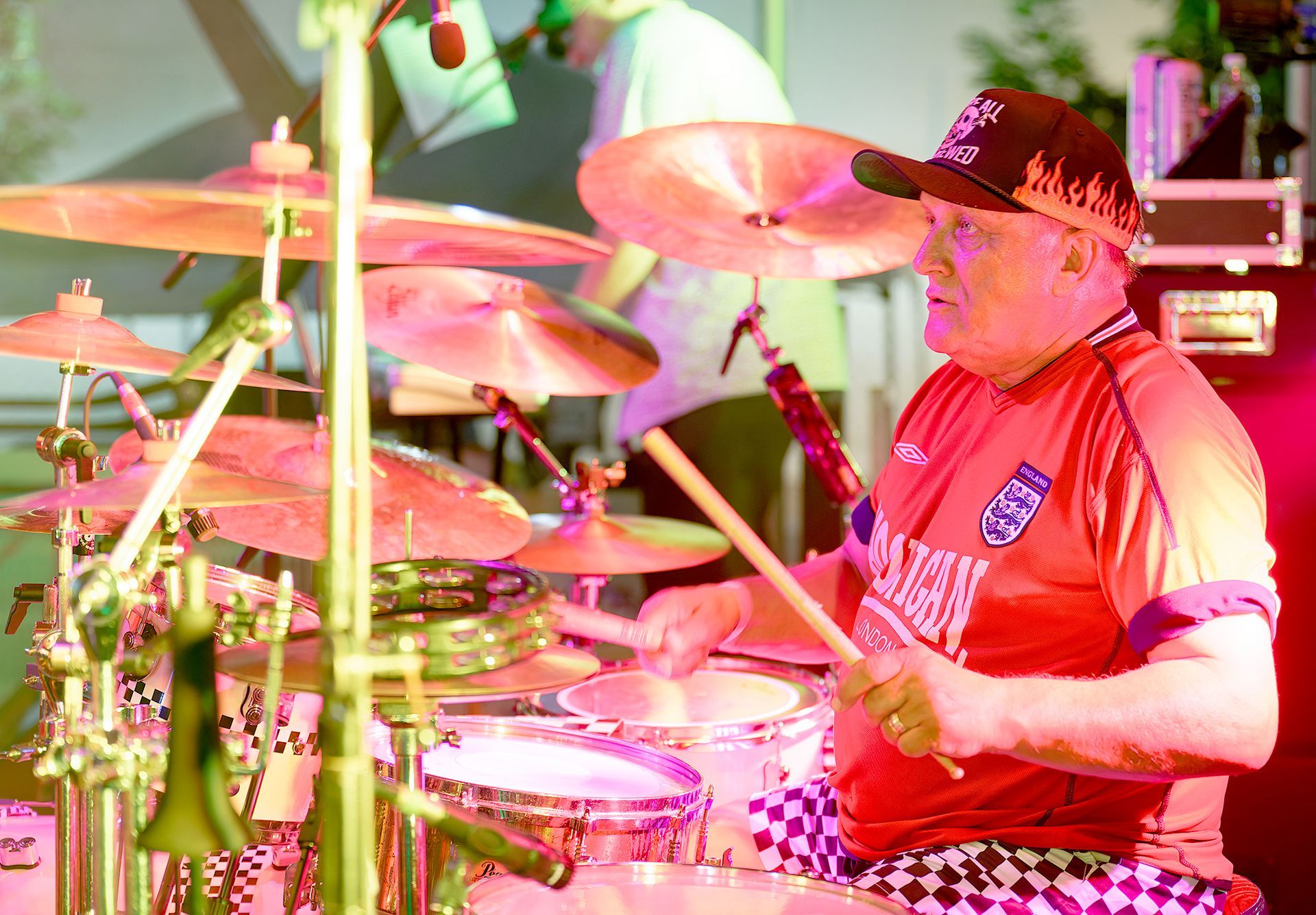 Drummer in red shirt and black hat playing a drum kit onstage under pink lighting