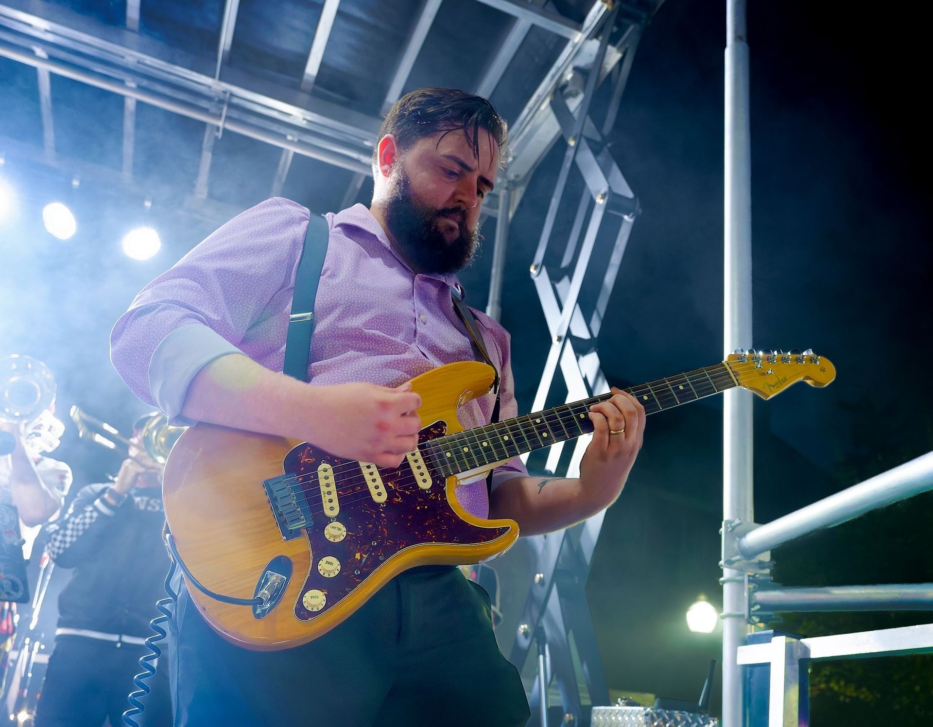 Musician playing yellow electric guitar on a brightly lit stage with blue lights and metal rigging