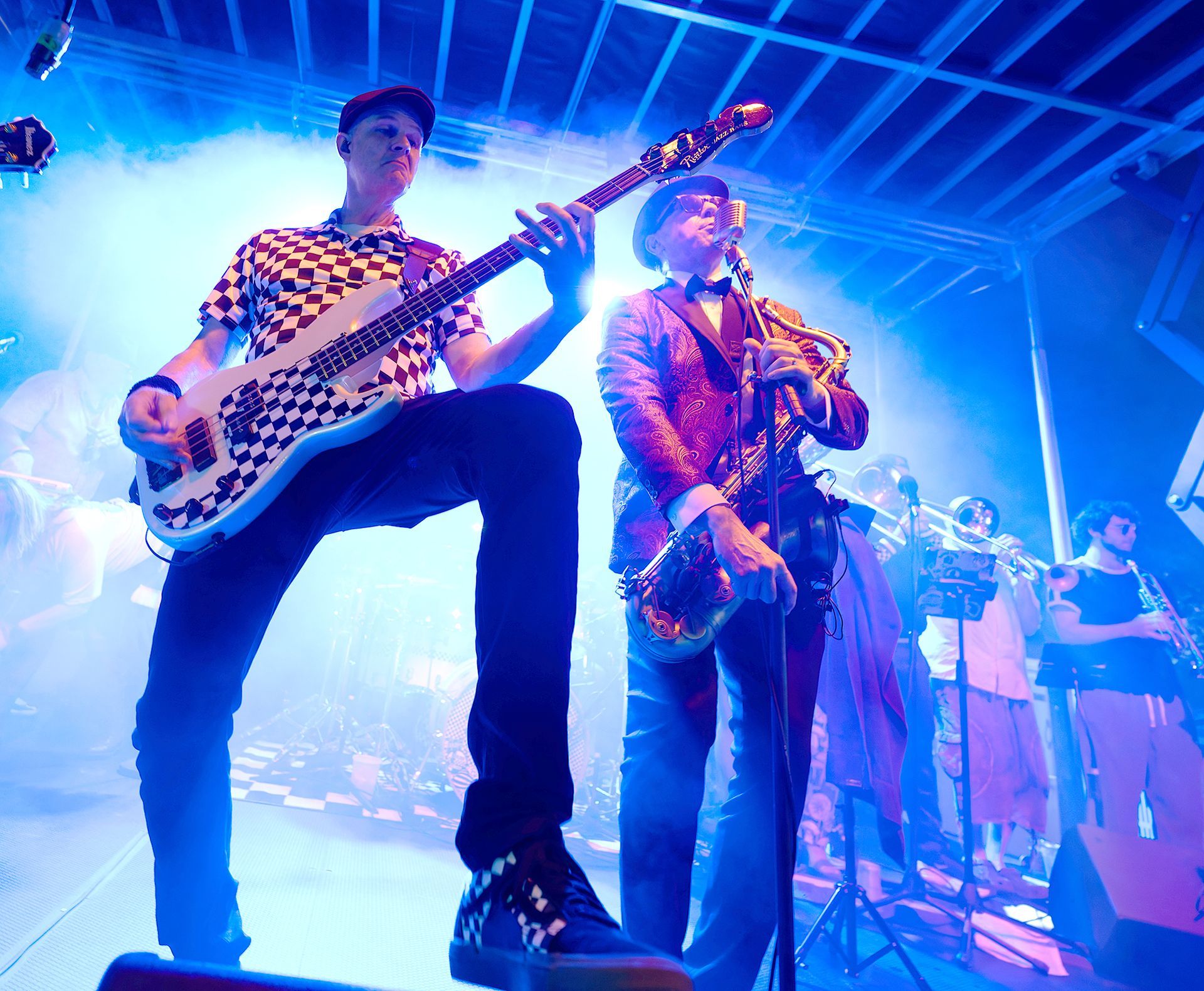Band performing on a blue-lit stage, with a guitarist in the foreground and horns in the background.