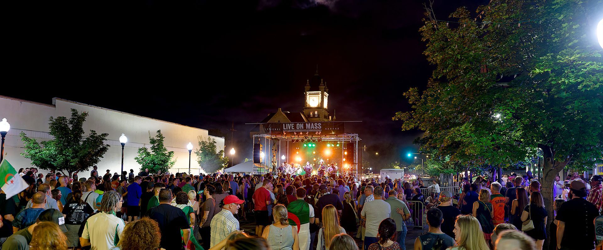 Crowd gathered at a brightly lit nighttime festival with a decorated stage and trees nearby