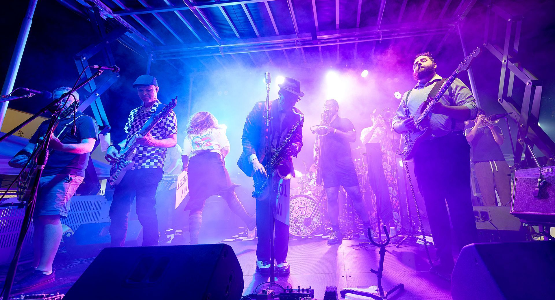 Band performing onstage under vivid purple and blue lights, with guitars and drums visible.
