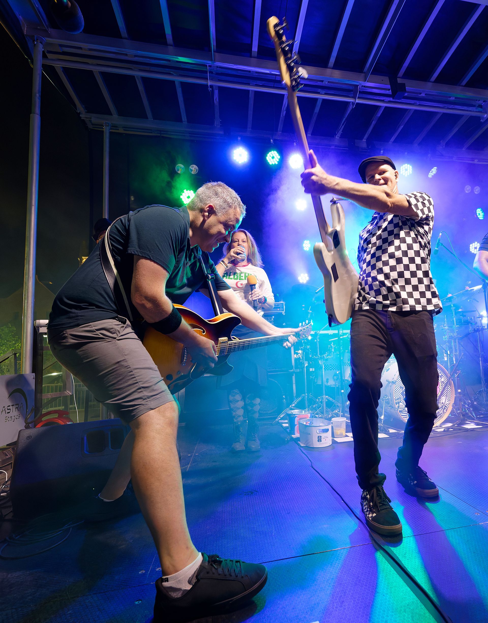 Two musicians performing on a blue-lit stage, one crouching with a guitar and one holding a raised guitar mic stand.