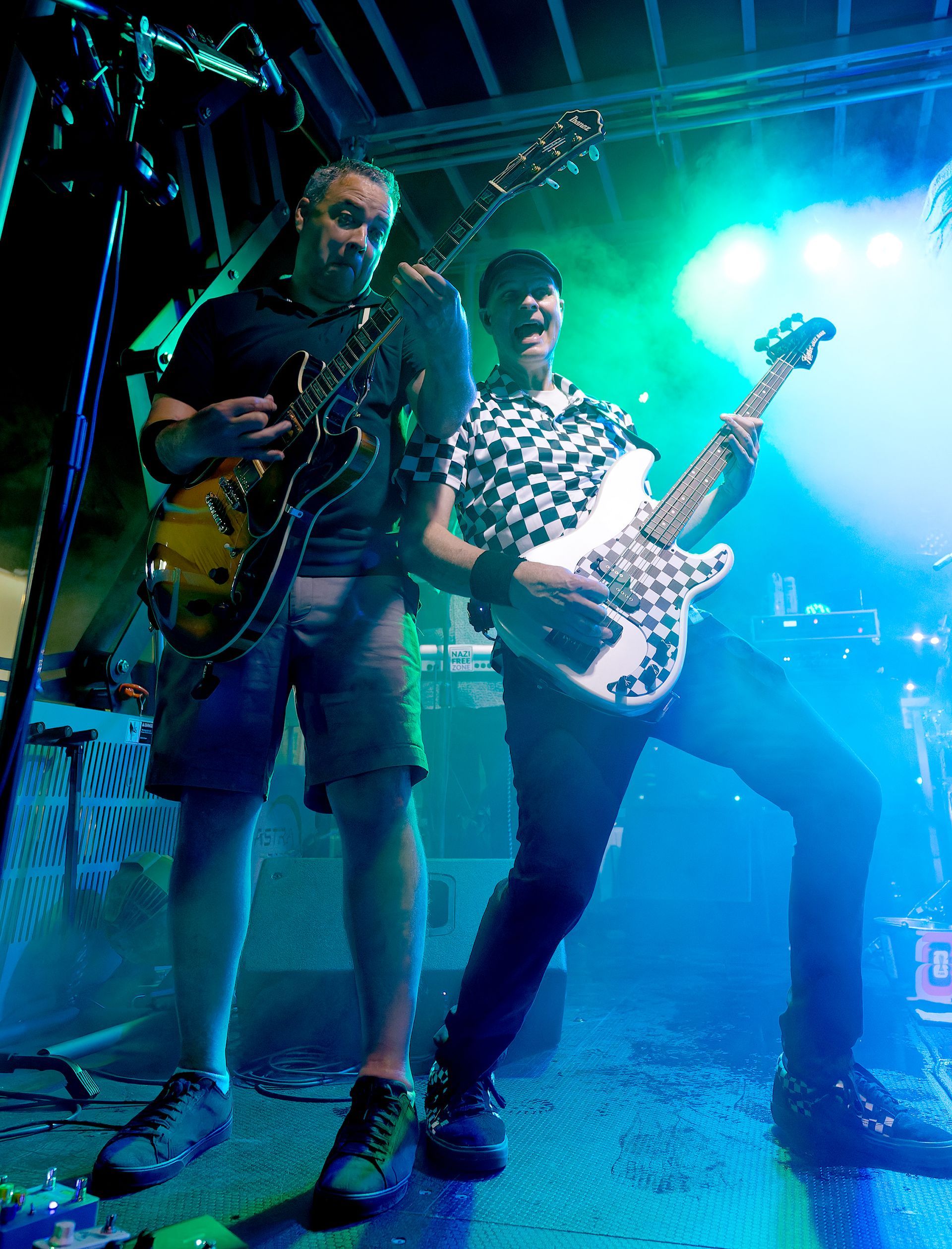 Two musicians play guitars on a brightly lit stage with blue and green lights.
