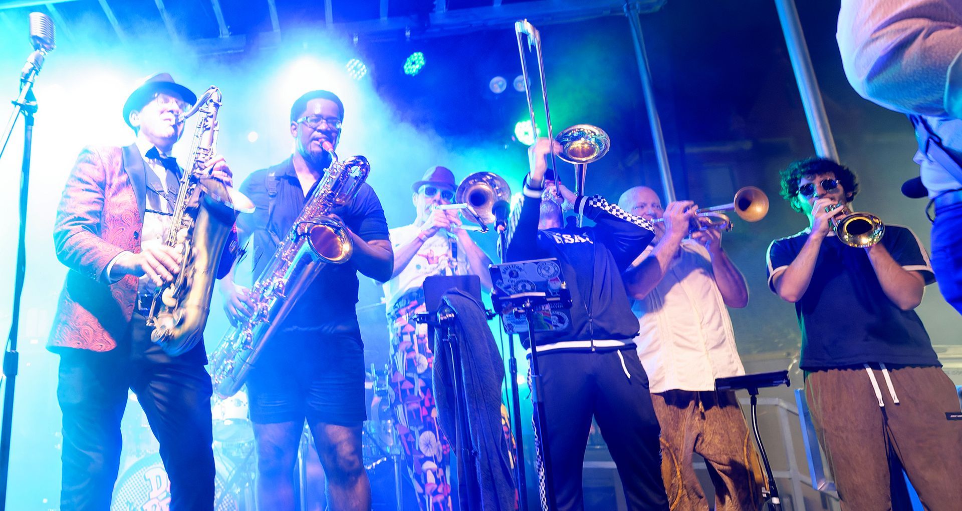 Jazz band performing on a brightly lit stage with saxophone, trumpet, trombone, and percussion