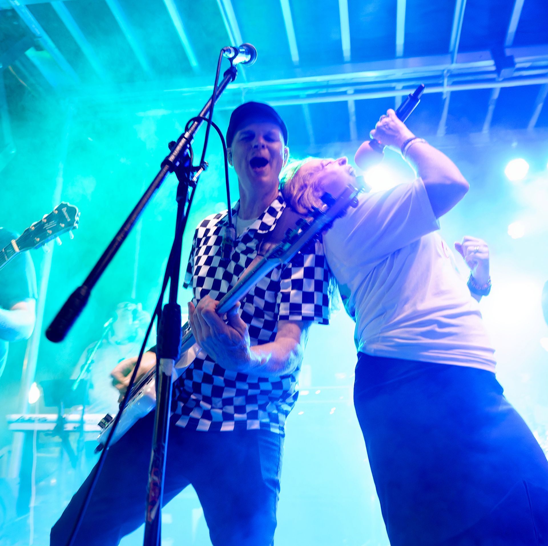 Two musicians performing on stage under blue lights, one singing into a mic and one playing guitar.