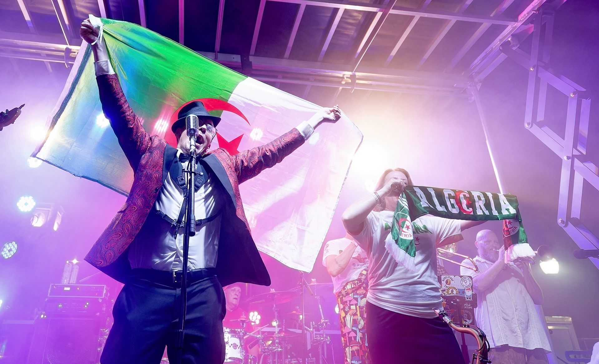 Performer onstage waving a flag under purple lights, with a crowd and banner behind them.