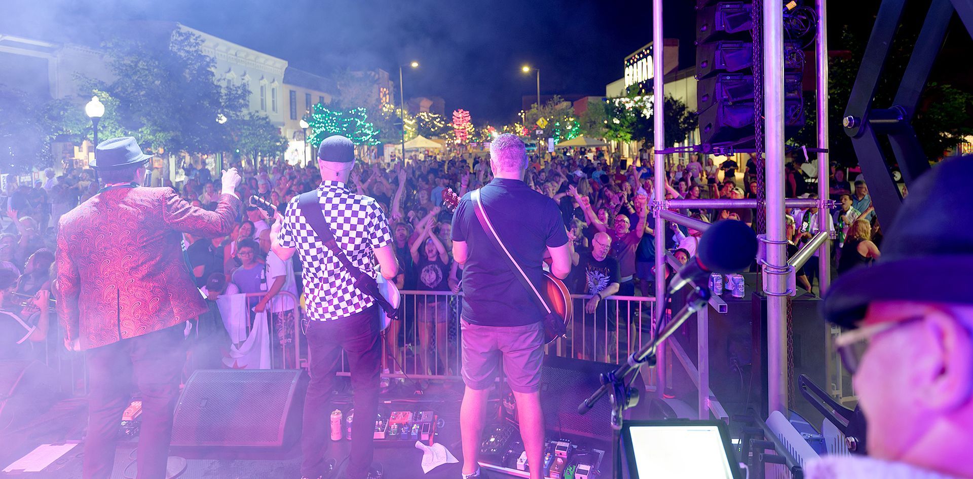 Band performing onstage at a night concert, seen from behind with a crowd and purple lighting