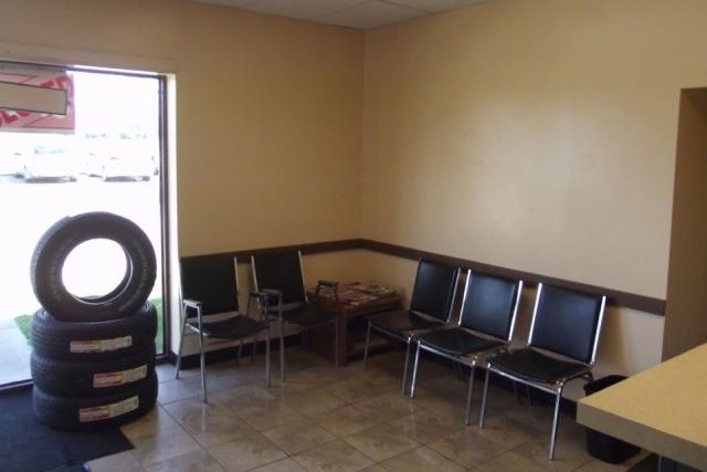 Empty waiting room with metal chairs, a small table, tan walls, and a tire display by the window.