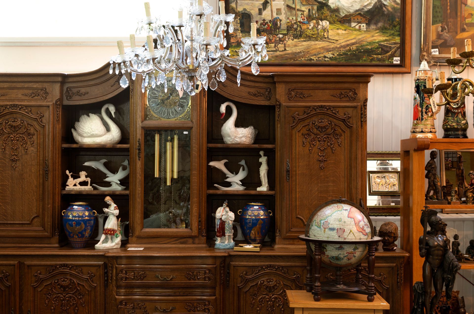 Antique wooden cabinet with swan figurines, a globe, and a chandelier in a shop setting.